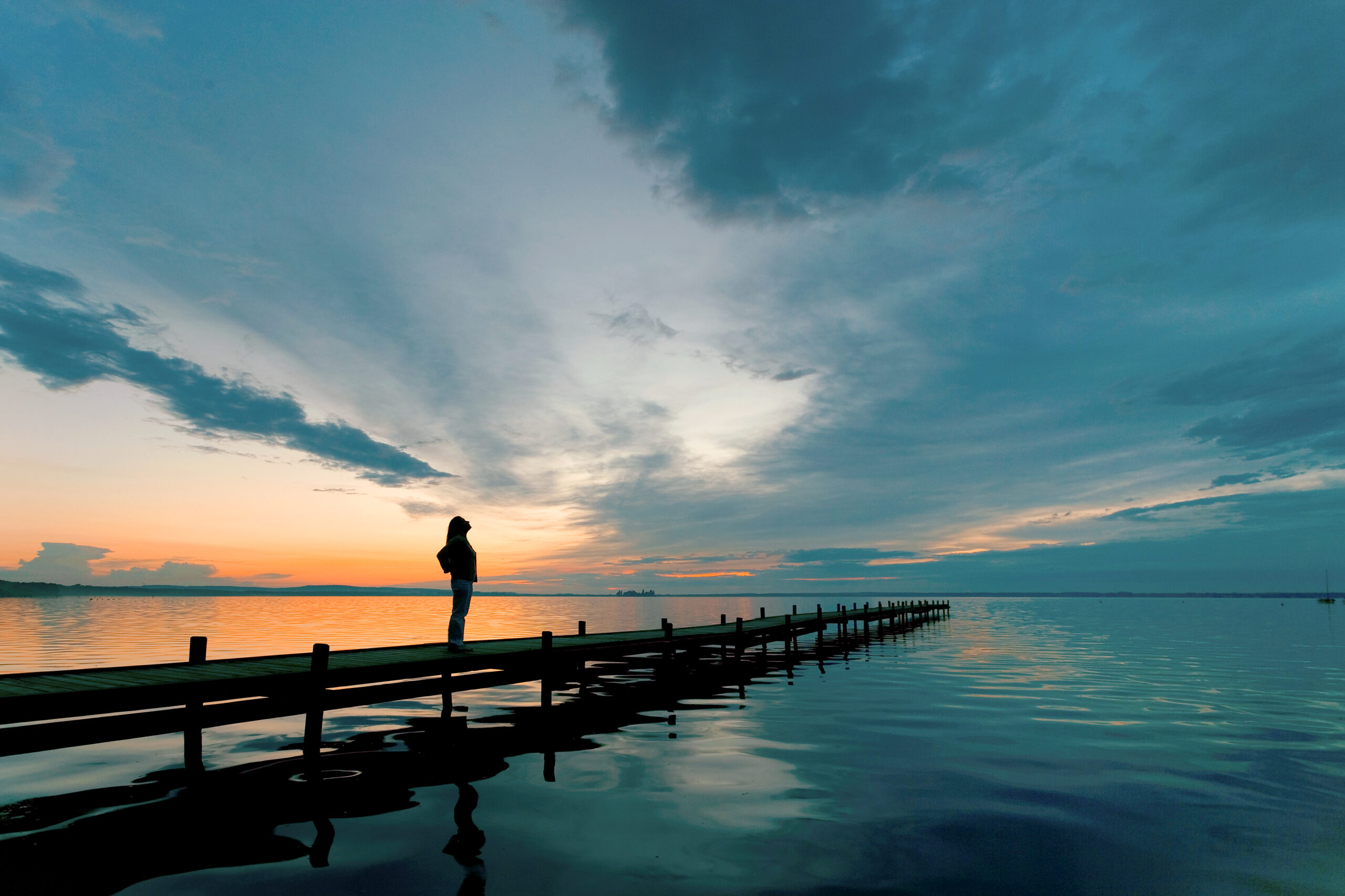 Young woman standing lakeside on jetty having a look at magical cloudscape and sunset colors.