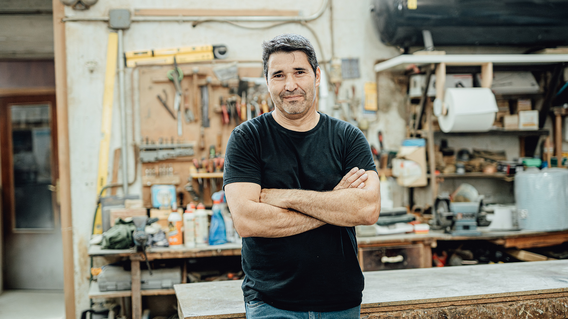 Confident middle-aged craftsman standing in his indoor workshop, smiling and working on a woodworking project with various tools and equipment on his workbench