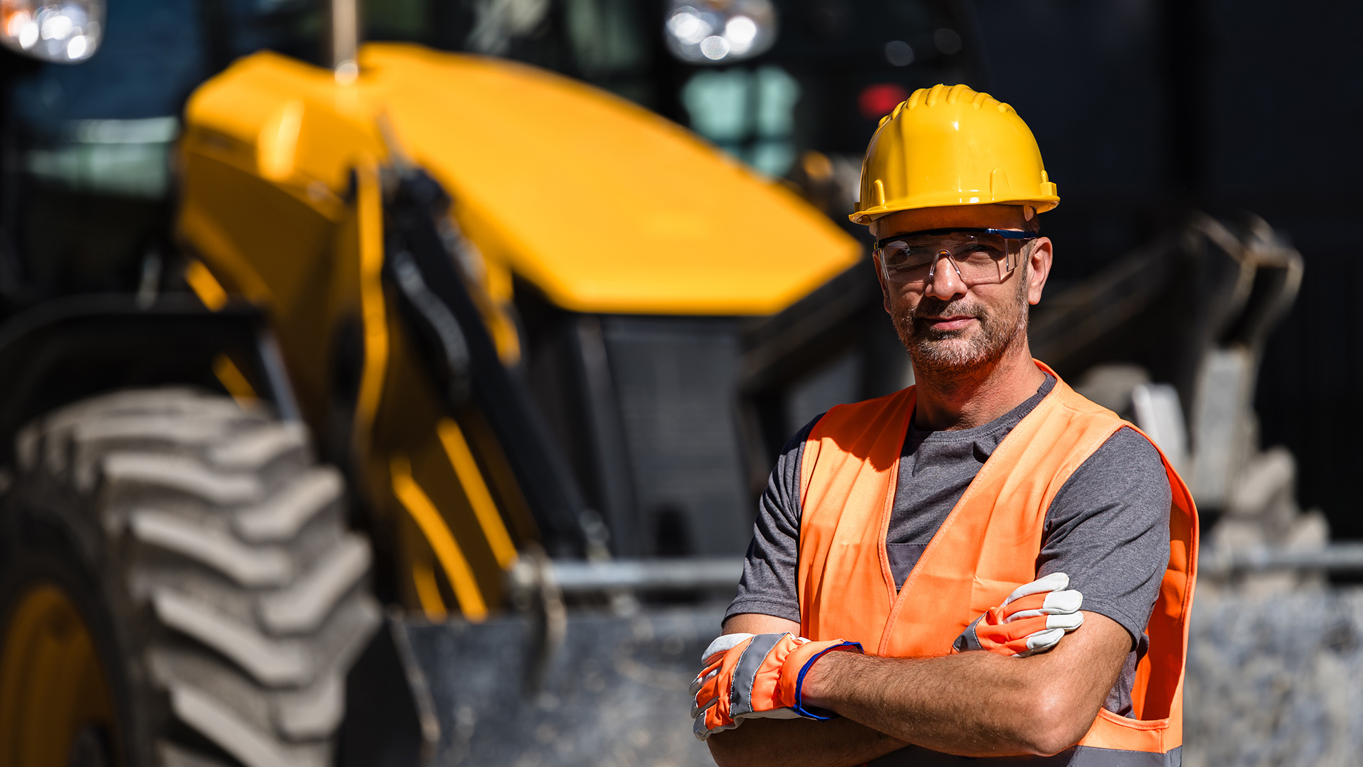 Construction worker in safety gear stands confidently near a yellow heavy machinery during daylight hours at a job site