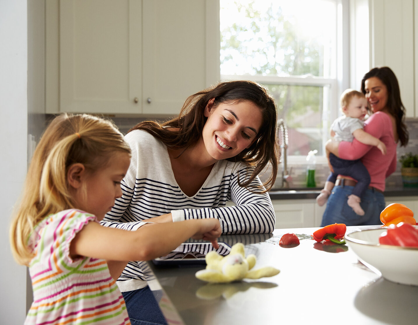 Girl uses tablet in kitchen with mum, other mum holding baby