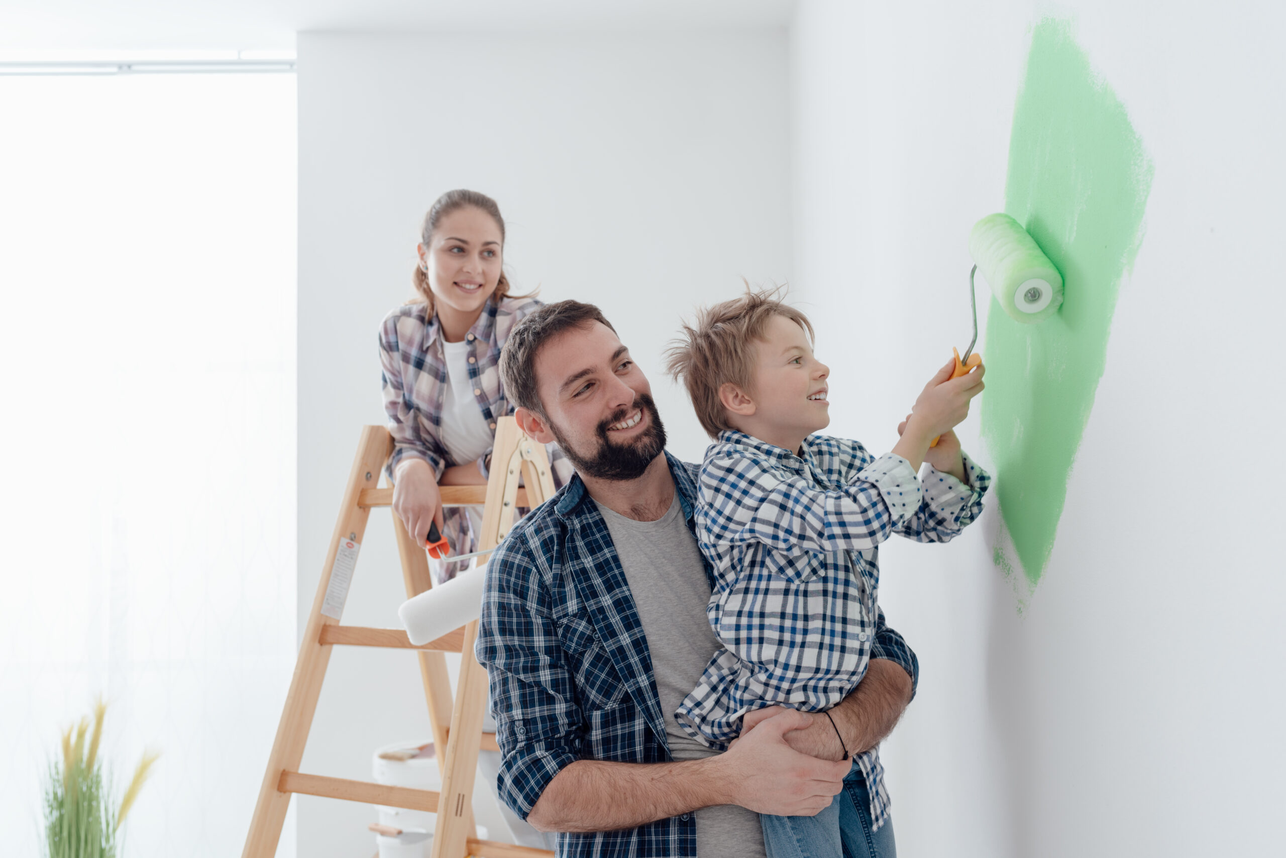 Happy young family renovating their home, the father is holding his son and he is helping him to paint a wall with a paint roller, the mother is standing on the ladder and smiling