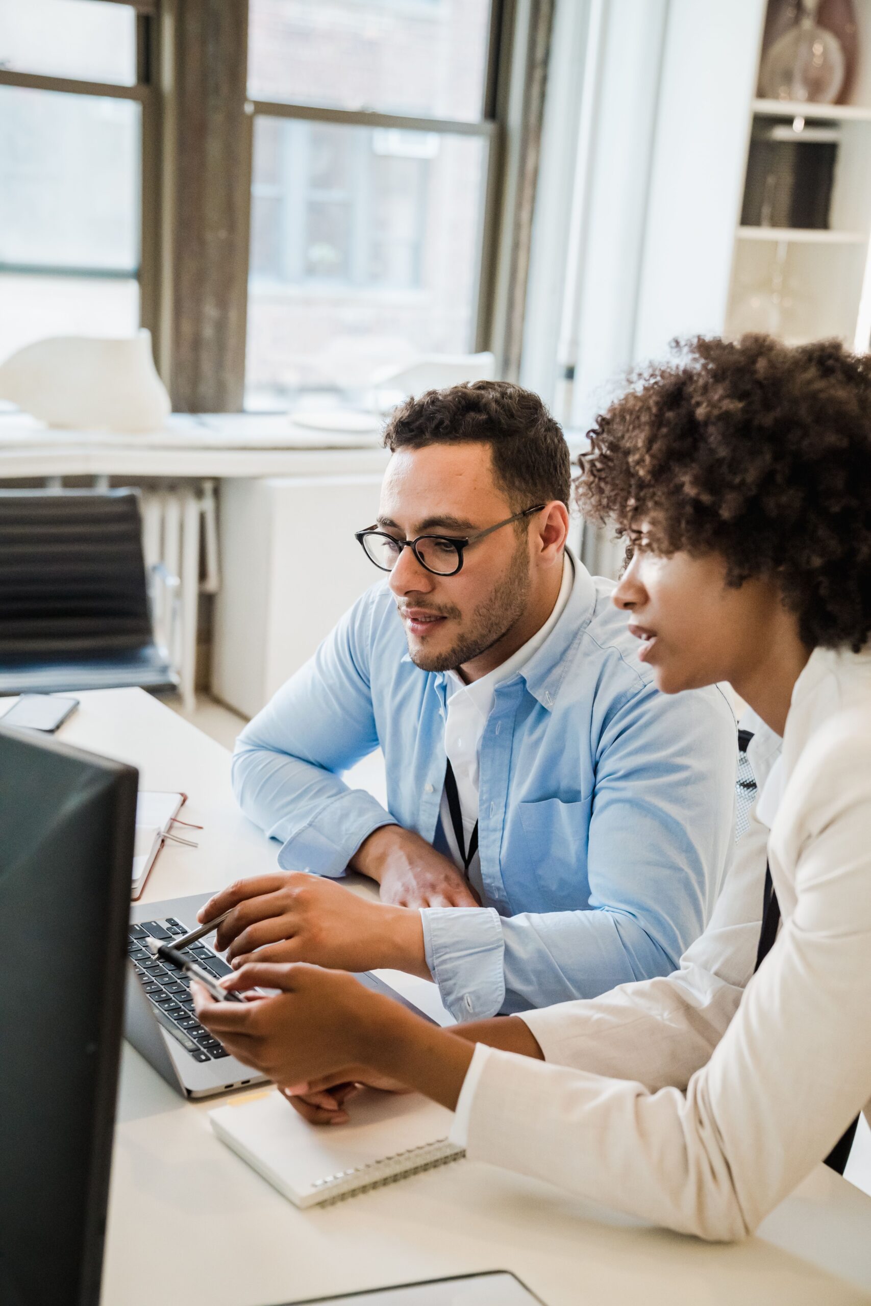 Two colleagues sitting at a desk together. One colleague has a pen in his hand and is pointing at the screen. The other colleague is offering advice.